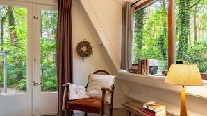 A cozy room with a wooden chair, a small table with books, a lamp, and a view of greenery through a window. - Boshuisje Rekem Two-Bedroom Houses (Lanaken)