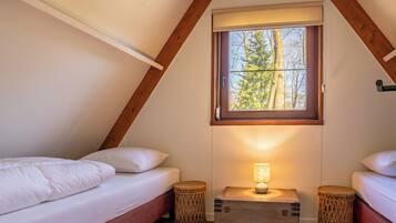 A cozy bedroom with two beds, a window with a view of trees, and a bedside lamp.