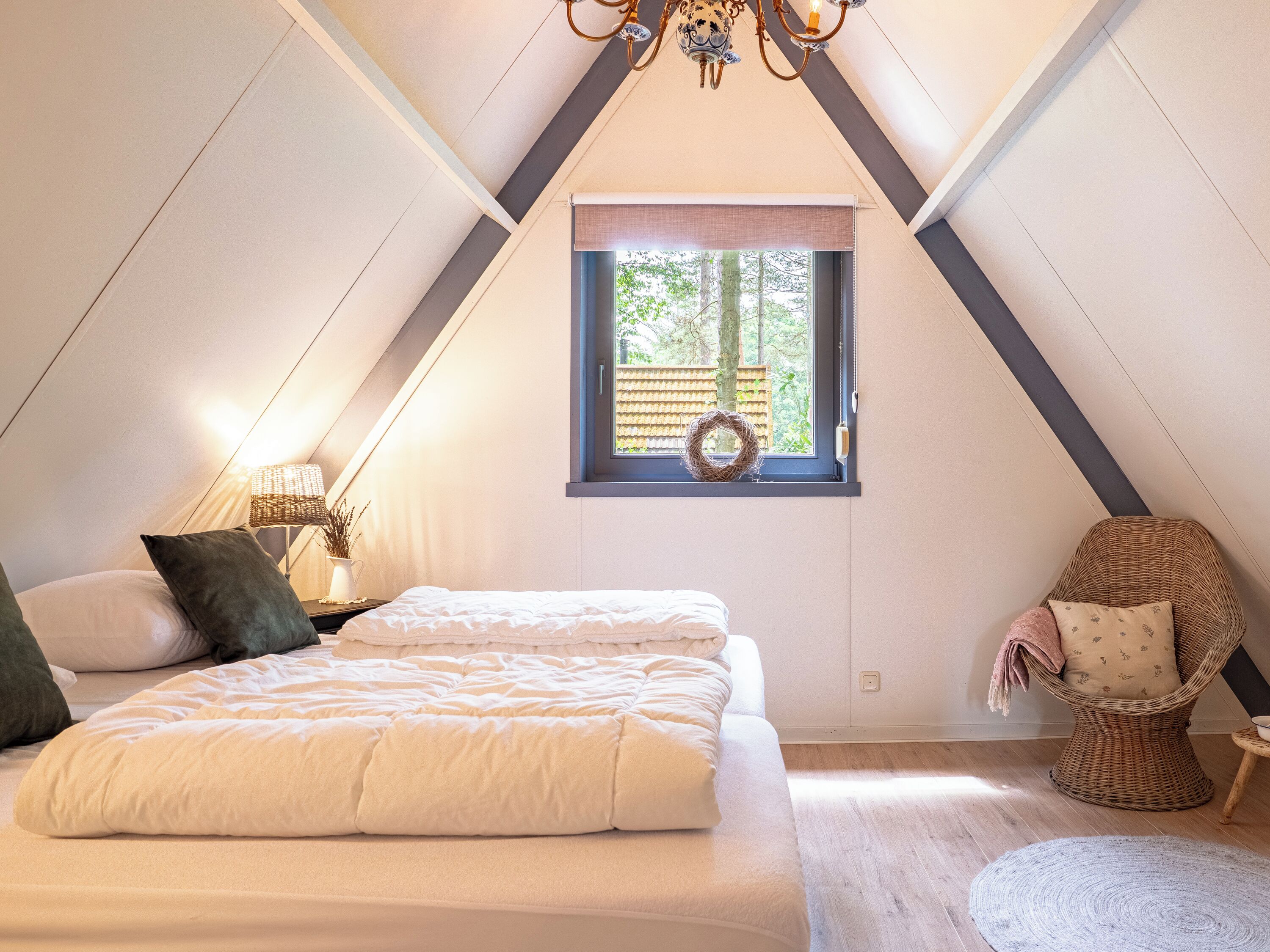 Ein Schlafzimmer mit einem Bett, einem Nachttisch, einer Lampe, einem Korbstuhl, einem Fenster mit einem Kranz und einer schrägen Decke.