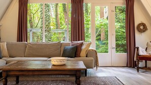 A cozy room with a sofa, a wooden table, and a view of greenery outside. - Boshuisje Rekem Two-Bedroom Houses (Lanaken)