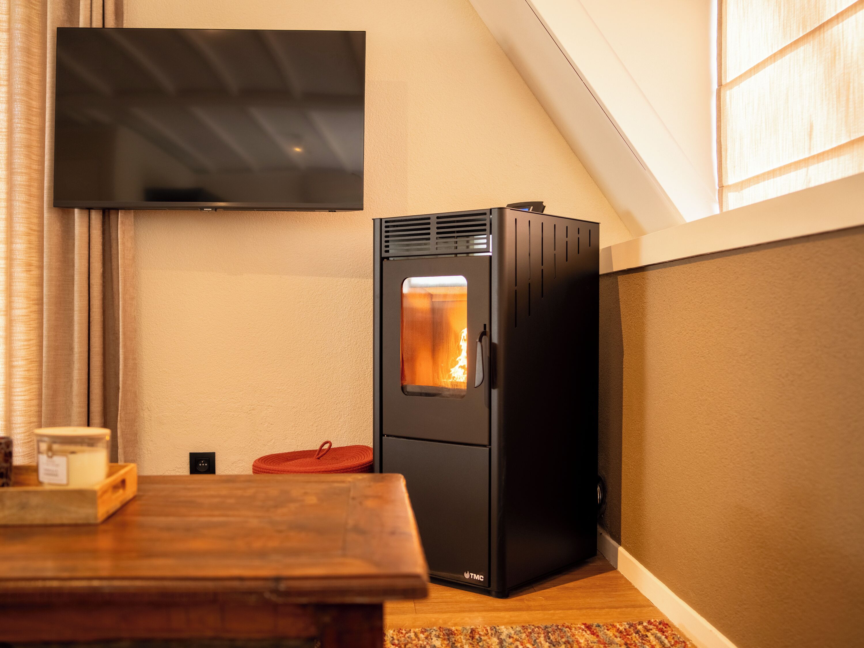 Una stanza con una stufa a legna in legno nero, una TV a schermo piatto montata a parete e un tavolo di legno con un sottotavolo rosso.