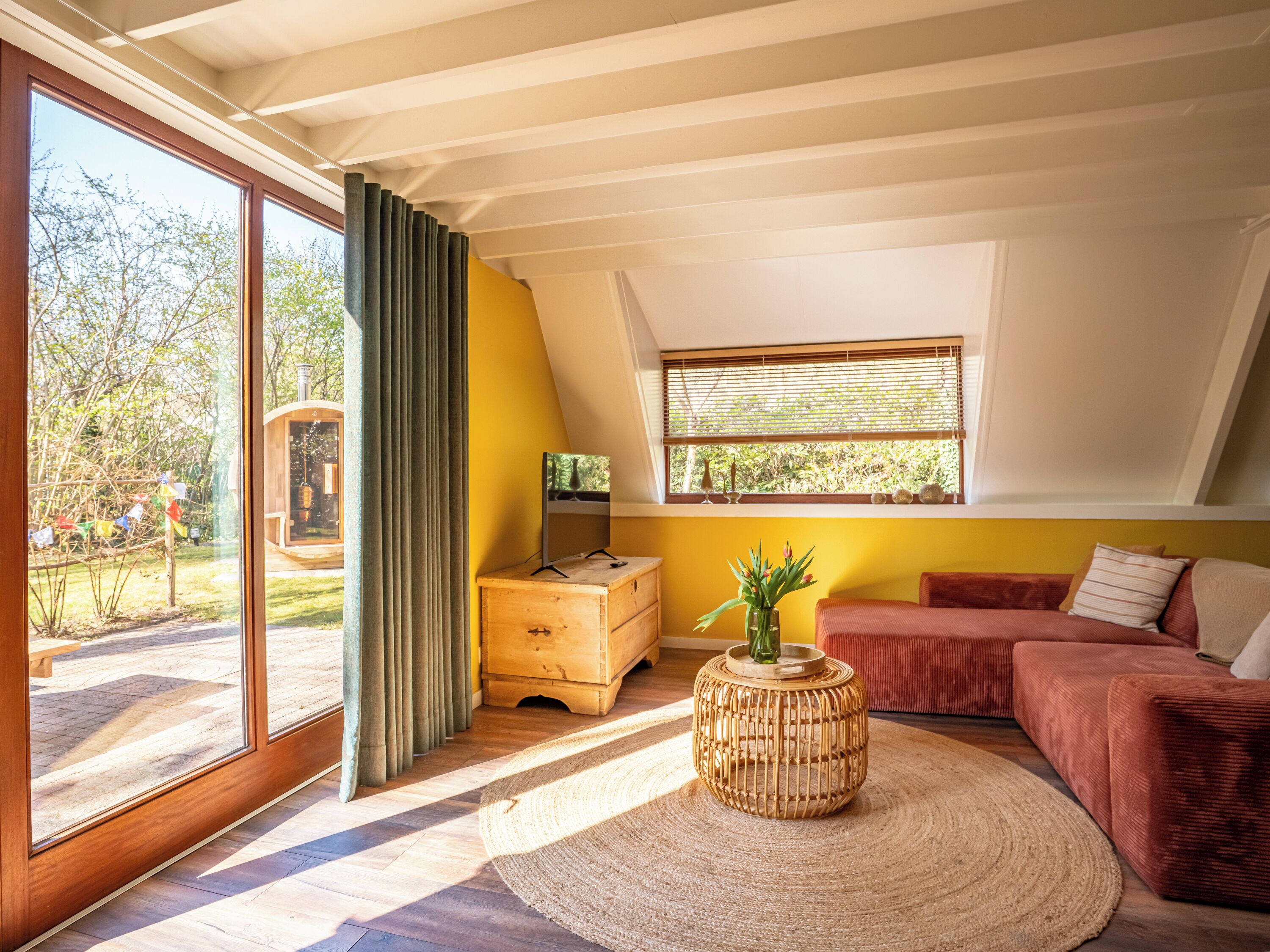 A modern living room with a large sliding glass door, a wooden bench outside, a red sofa, a small wooden table, and a window with blinds.