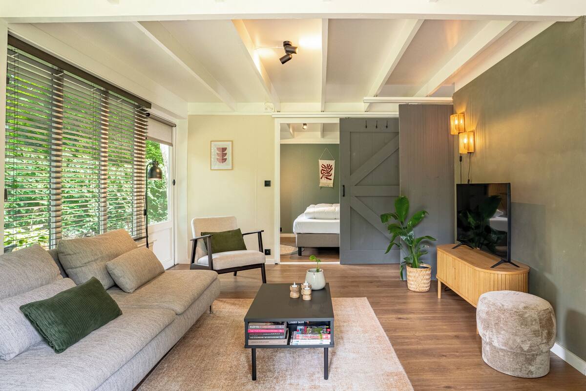 A modern living room with a sofa, coffee table, and a TV. There is a bed in the background and a door leading to another room.