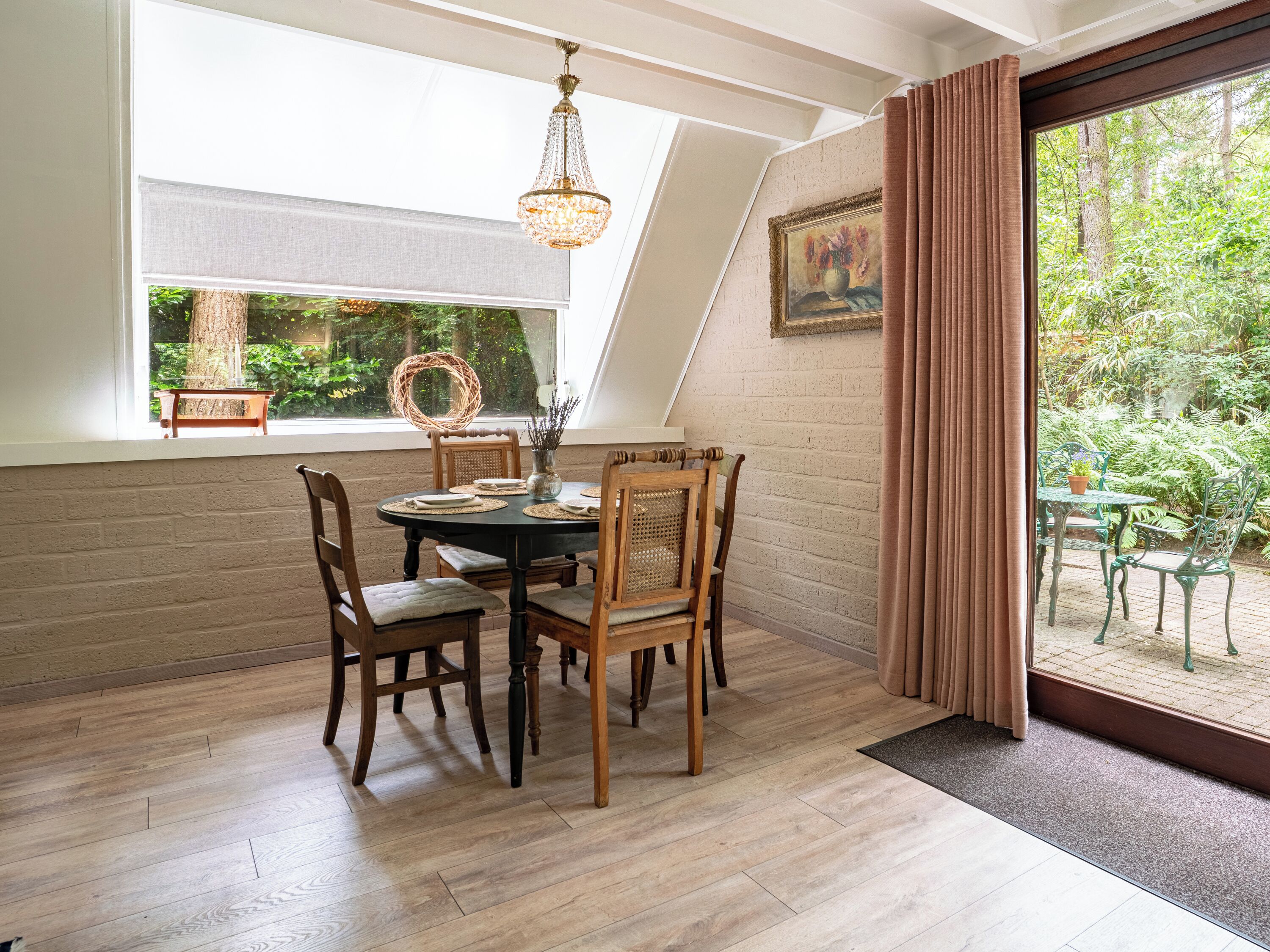 Een eethoek met een ronde tafel, houten stoelen, een kroonluchter en uitzicht op een tuin door een raam.