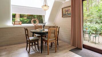 Een eethoek met een ronde tafel, houten stoelen, een kroonluchter en uitzicht op een tuin door een raam.