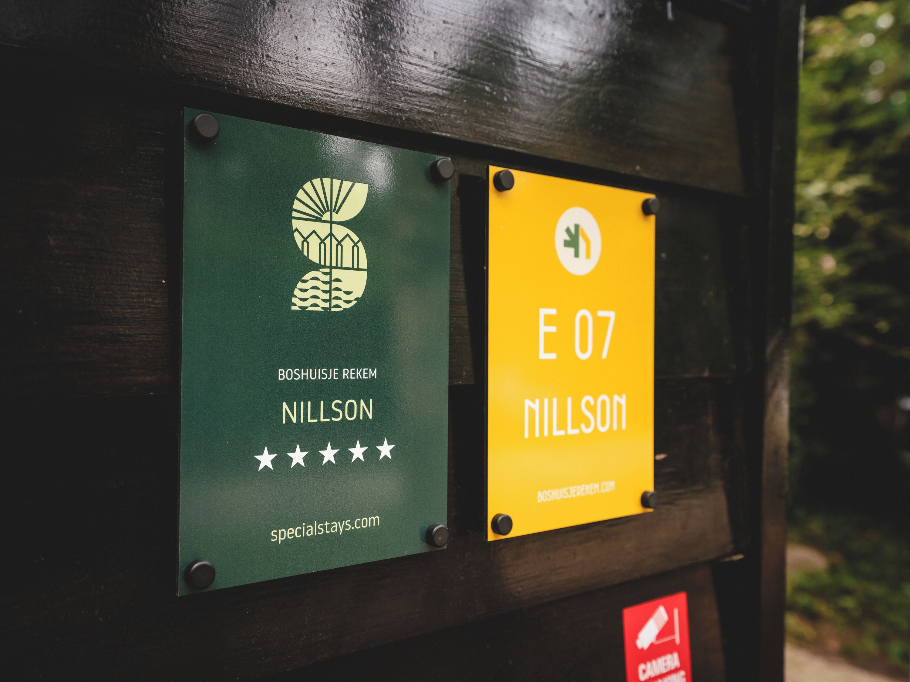 Two signs on a wooden structure: one green with a logo and text for 'Boshuisje Rekem Nillson' and a website, and one yellow with 'E 07 Nillson' and a website.