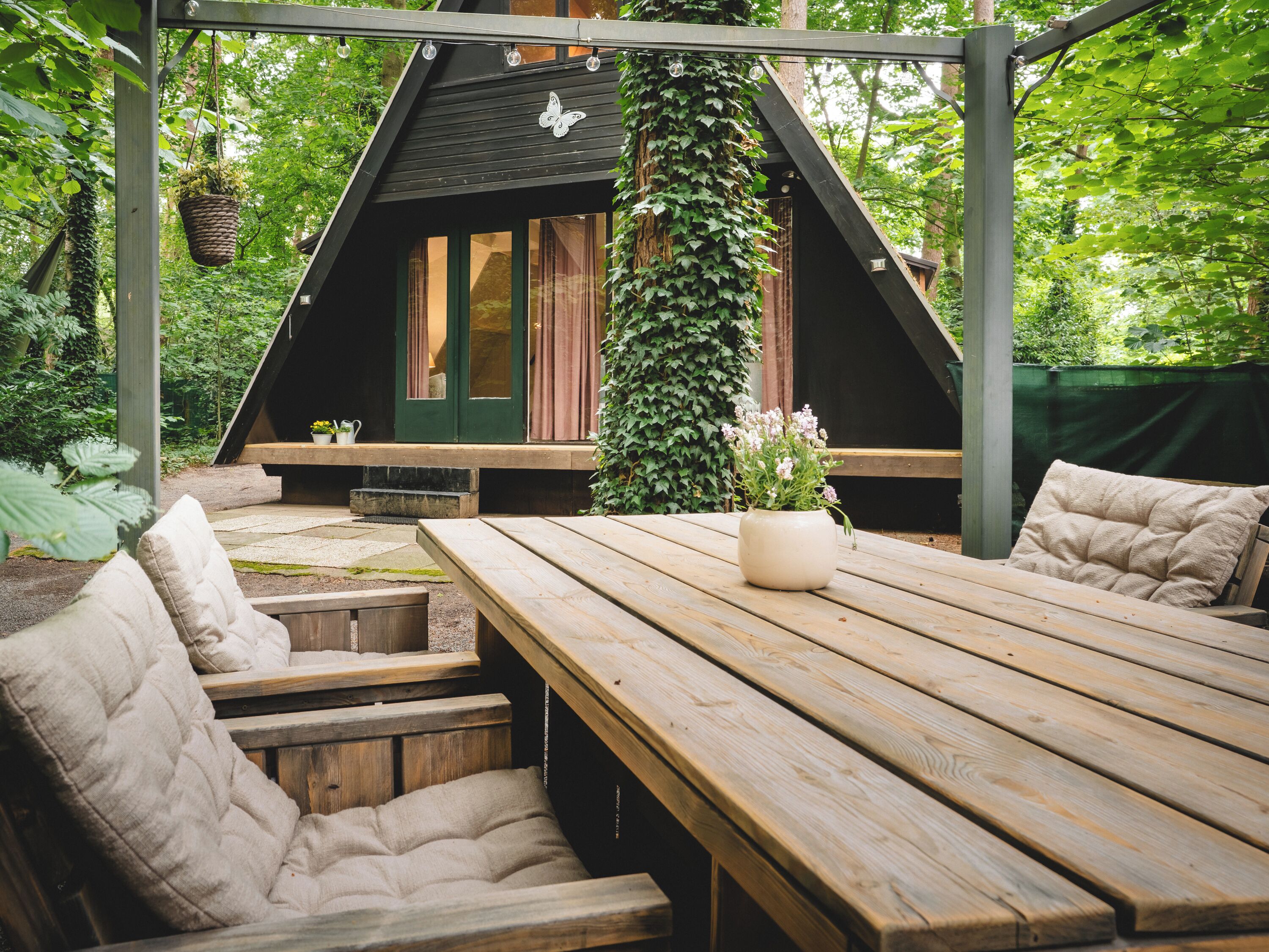 Una terraza de madera con una mesa de picnic y bancos, rodeada de follaje y una pequeña casa con techo inclinado.