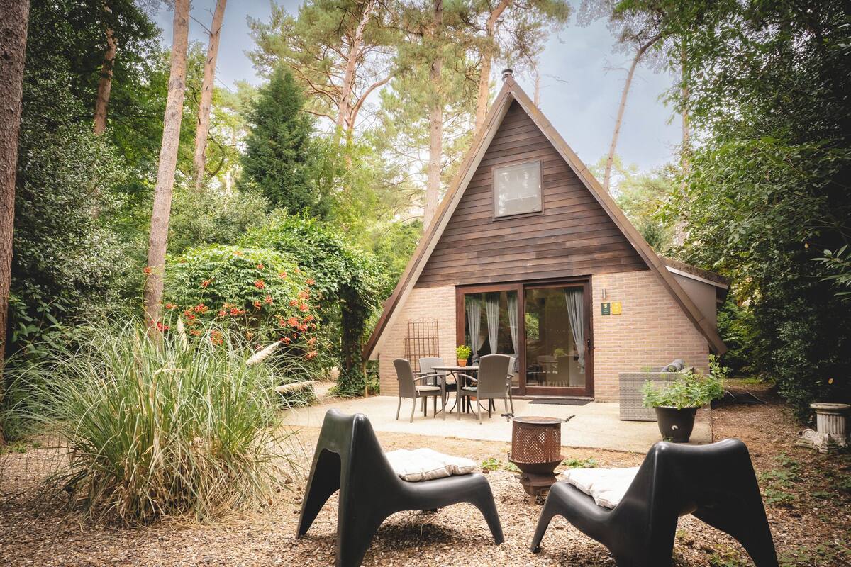 A small, wooden cabin with a triangular roof, surrounded by dense greenery and trees.