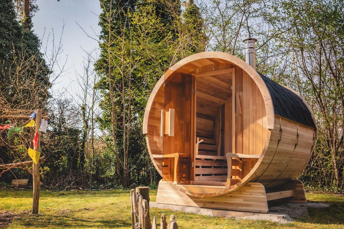 A wooden sauna structure with a curved roof and a chimney, situated in a grassy area with trees in the background.
