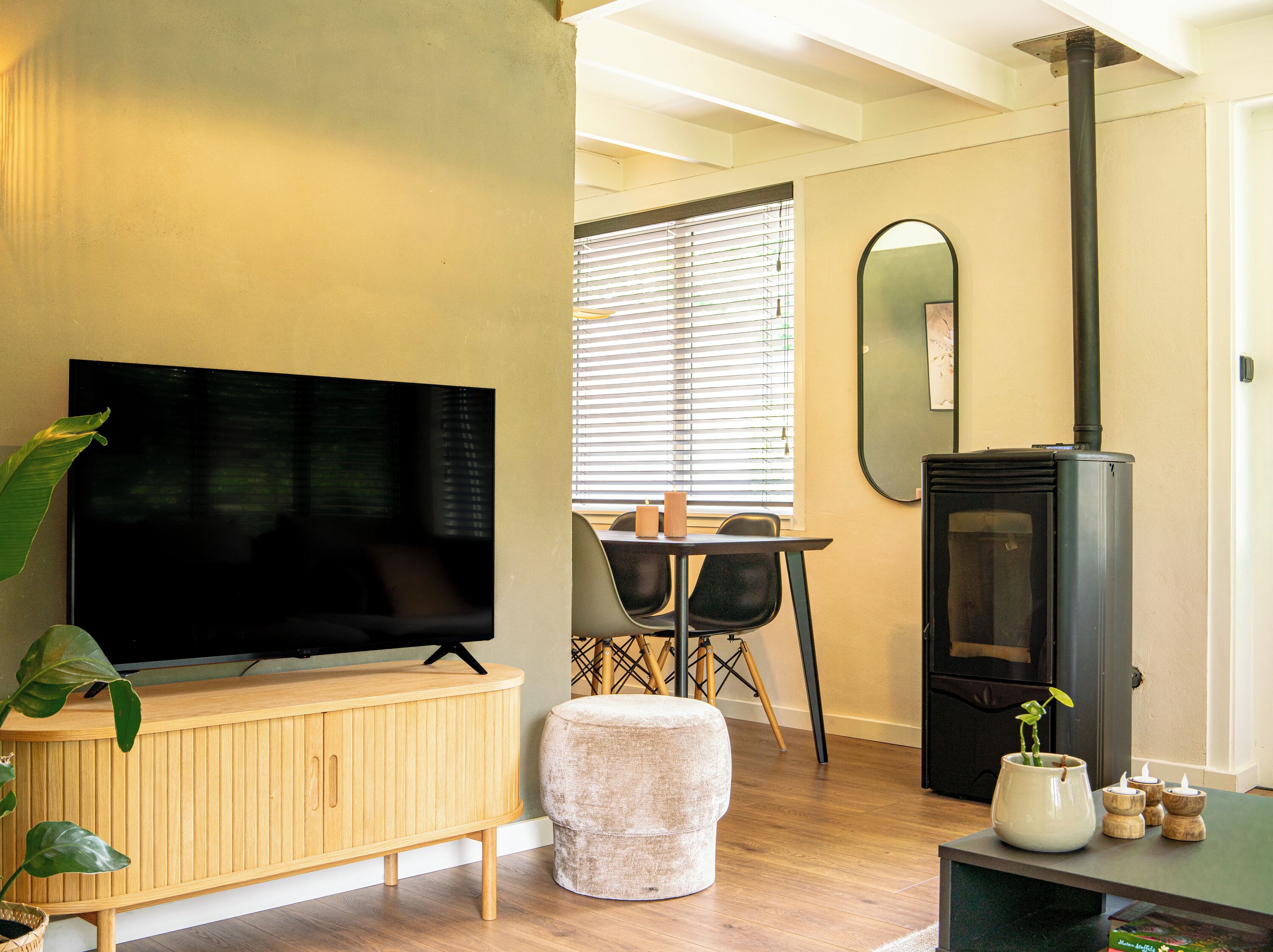 Una sala moderna con un televisor de pantalla plana, un mueble de madera, un reposapiés, una mesa de comedor, sillas y un estufa de leña negra.