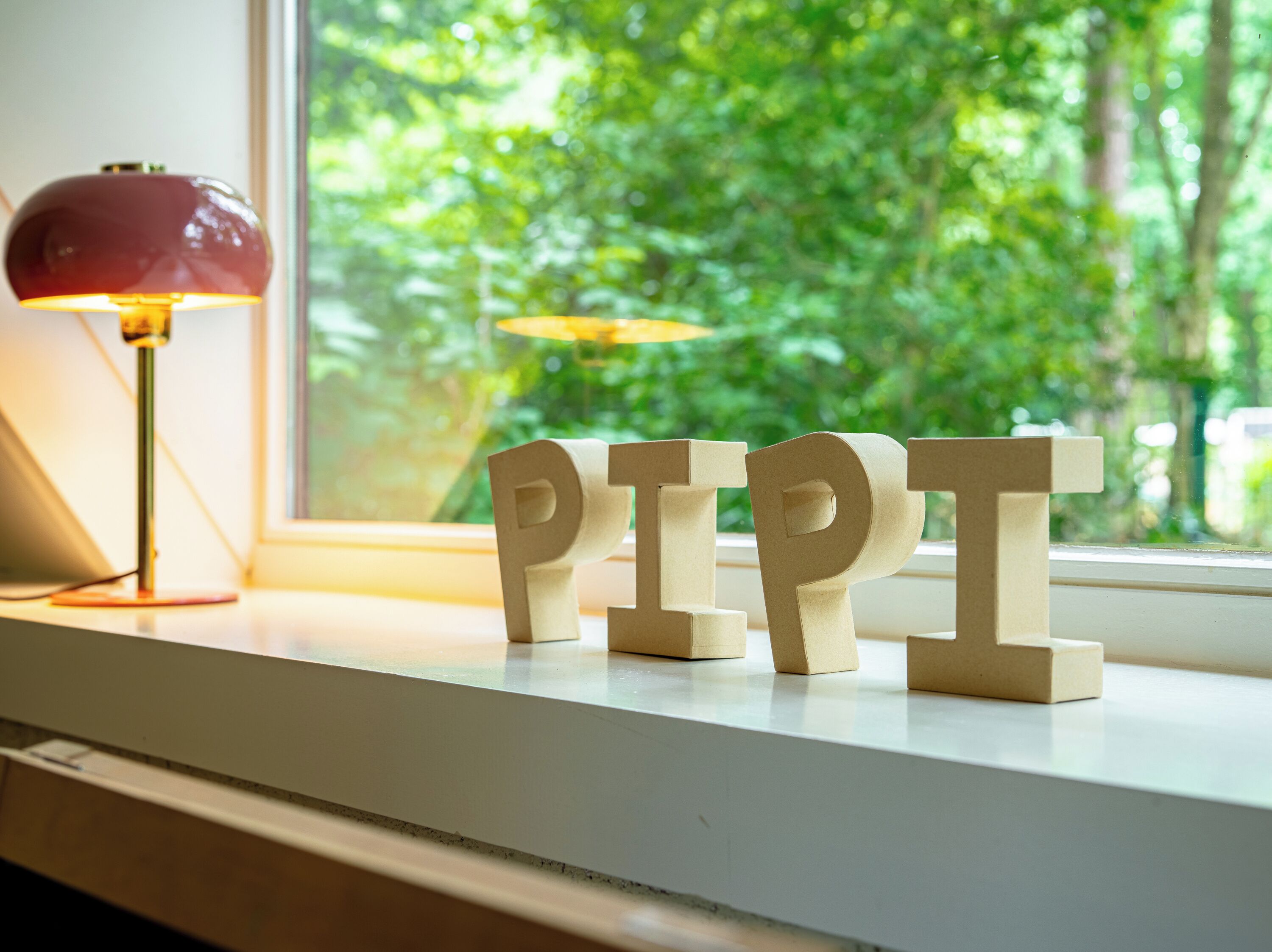 Træbogstaver, der staver "PIPT", på et vindueskar med en lampe og grønne planter udenfor.