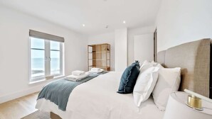 A modern bedroom with a large bed, a bedside table, a wooden shelf, and a window with a view.