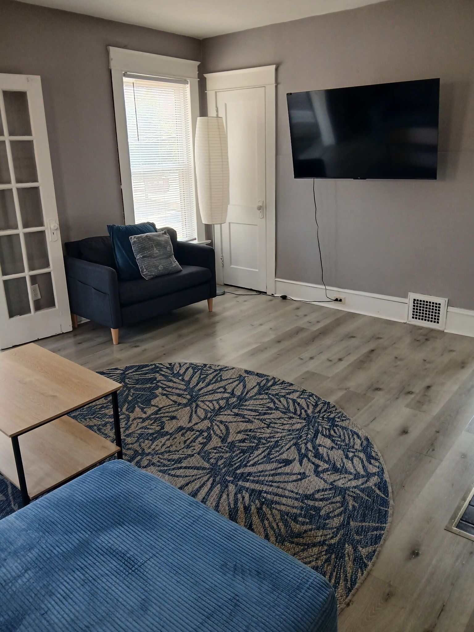 Un salon avec un téléviseur à écran plat fixé au mur, un canapé de couleur foncée agrémenté d’un plaid à motifs et une table basse en bois.
