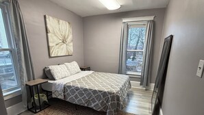 Une chambre à coucher avec un lit, une table de chevet, un tableau, une fenêtre avec des rideaux et un radiateur.
