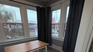 Une pièce avec une table et un banc en bois, de grandes fenêtres avec des rideaux et une vue sur les bâtiments à l’extérieur.