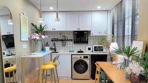 A compact kitchen with white cabinets, a built-in oven, microwave, and dishwasher. There are two yellow chairs and a wooden table with a vase of flowers. - Shanghai Hills&Zinn Changle road Homestay (Shanghai)