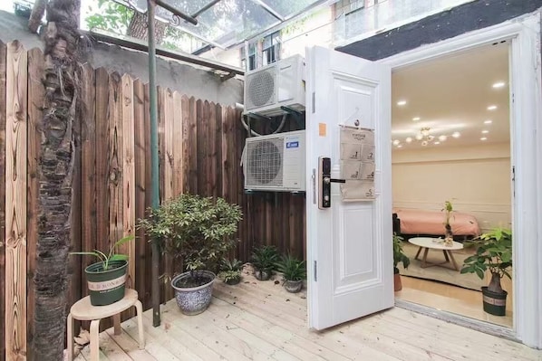 A room with a wooden floor, a white door, and a view into an adjacent room with a bed and a table. There are air conditioning units mounted on the wall and several potted plants.