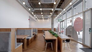 A modern meeting room with wooden tables and grey upholstered seating, large windows, and visible ductwork on the ceiling. - Cosy Stay in a Two Bed Flat (Milton Keynes)
