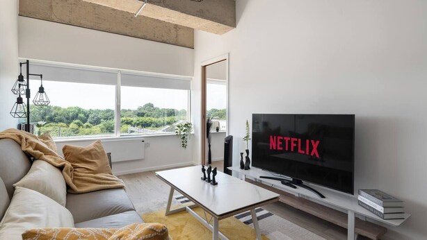 A modern living room with a large TV displaying 'NETFLIX', a sofa, a coffee table, and a view of greenery outside. - The Yellow Corporate Suite (Milton Keynes)
