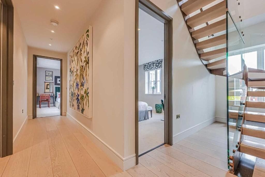 Un intérieur moderne avec un escalier en bois, un couloir menant à une pièce où se trouve un lit, et un grand tableau abstrait accroché au mur.