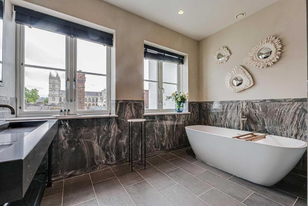 A modern bathroom with a freestanding bathtub, large windows with black blinds, and decorative wall mirrors.