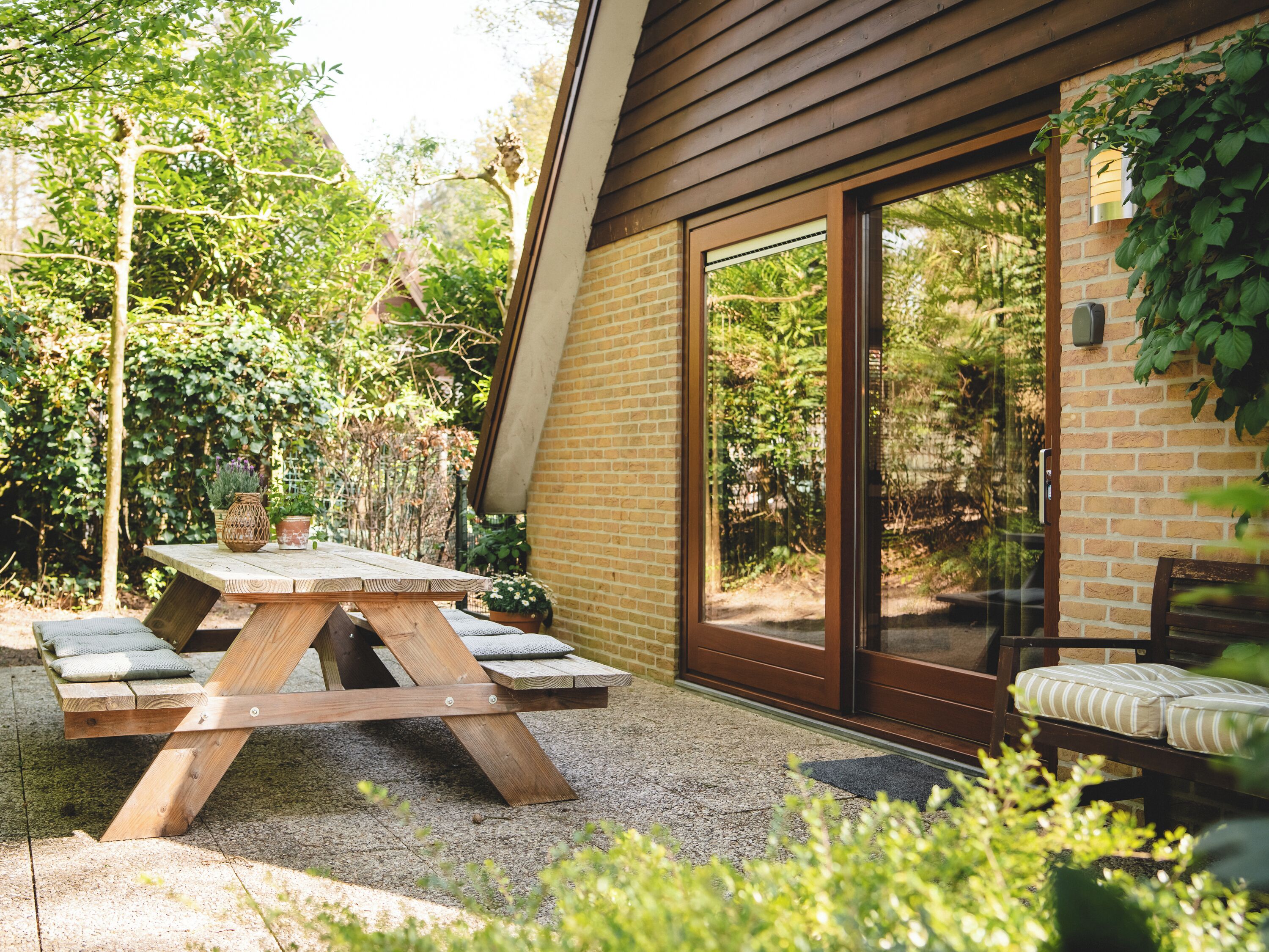 Een houten picknicktafel met kussens, een bakstenen muur en een glazen schuifdeur naar een tuin.