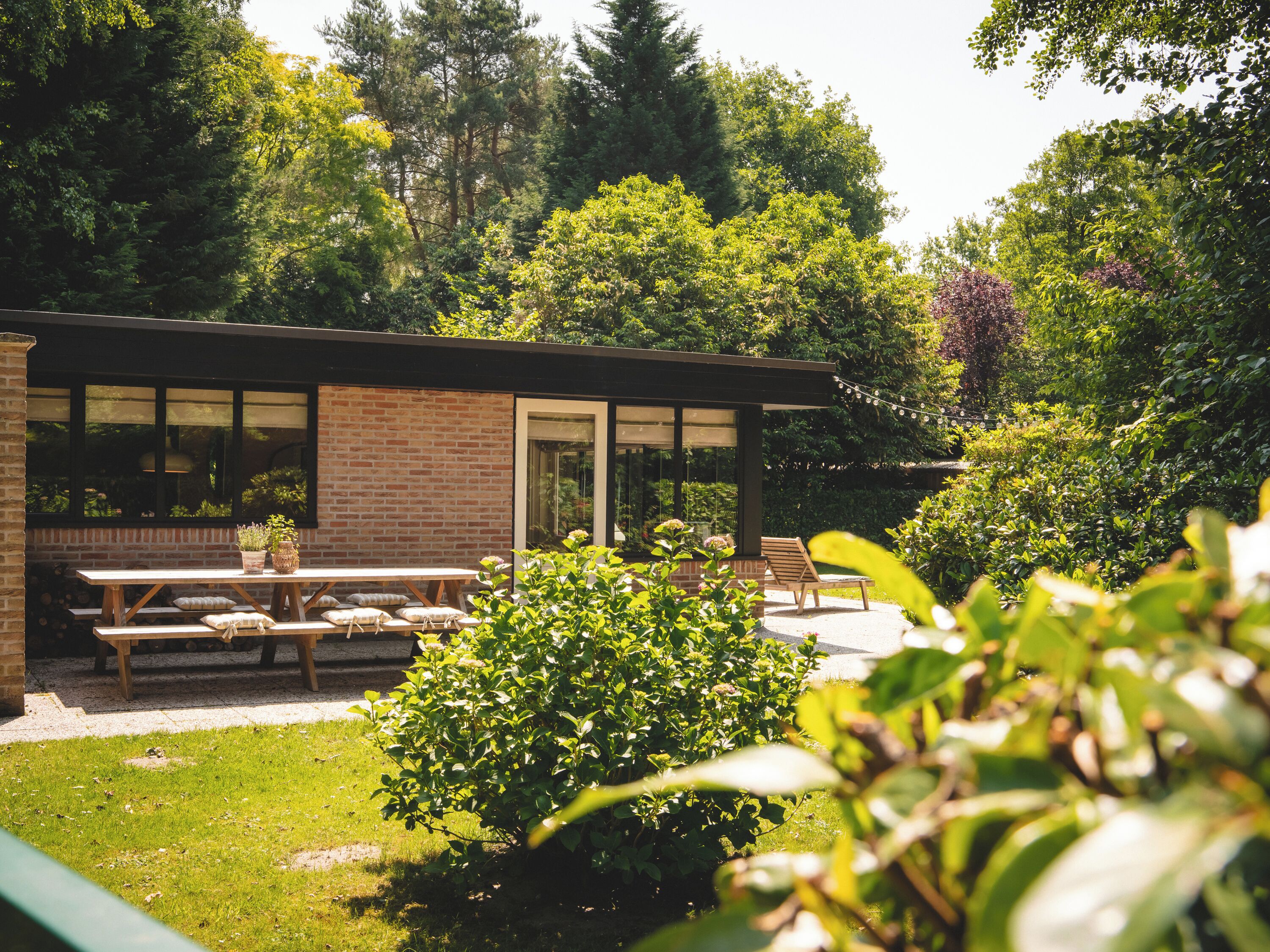 A modern building with a brick exterior and large windows, surrounded by lush greenery and a patio area with benches and a table.