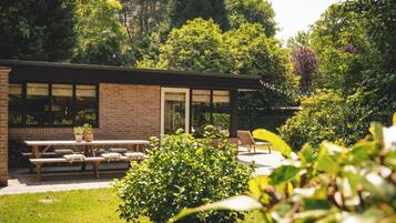 A modern building with a brick exterior and large windows, surrounded by lush greenery and a patio area with benches and a table.