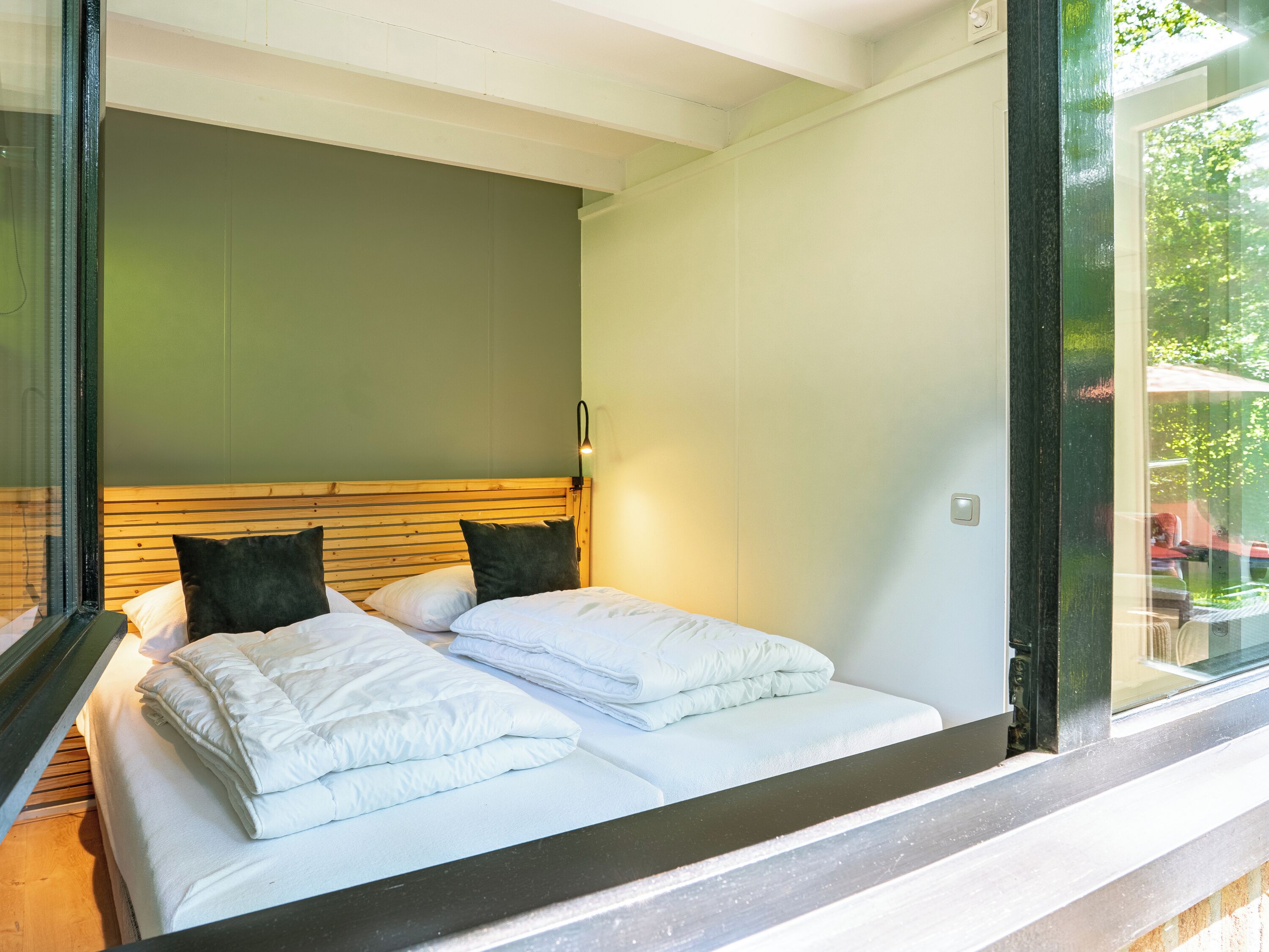 Ein modernes Schlafzimmer mit einem Holzkopfteil, zwei Betten mit weißen Bettwäsche und einem großen Fenster mit Blick auf Grünflächen.