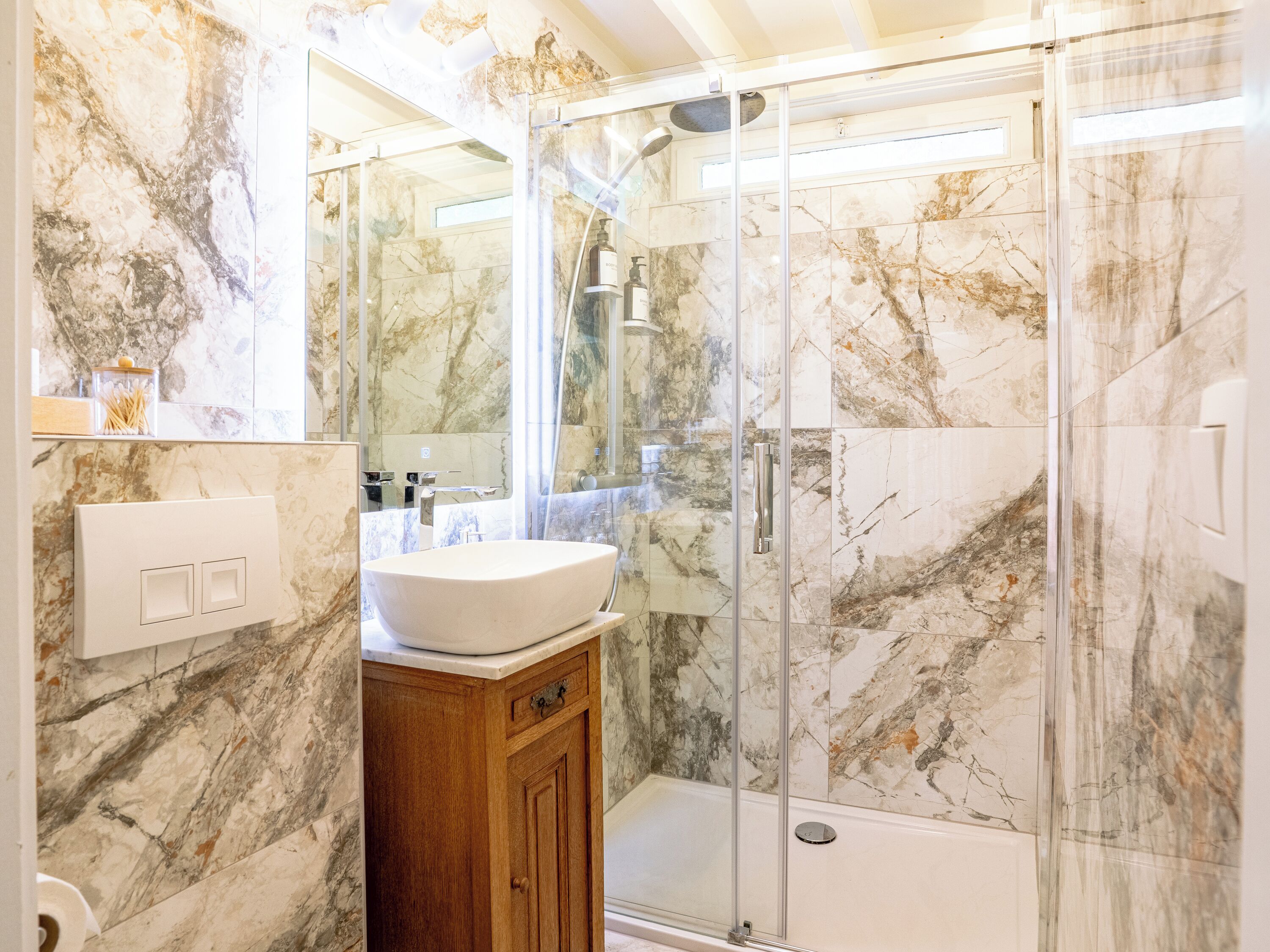 A bathroom with a glass-enclosed shower, marble walls, and a wooden vanity.