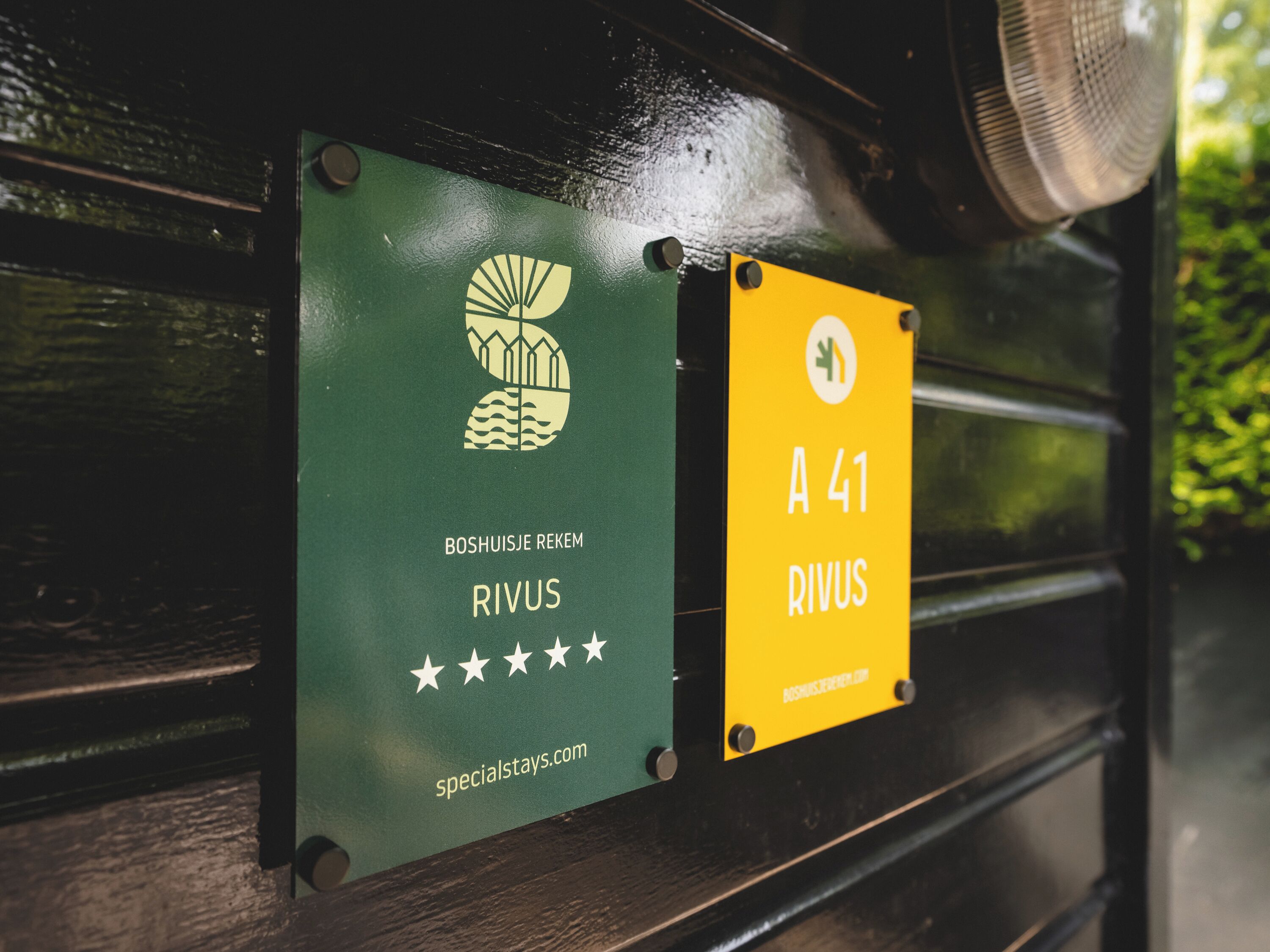 Two signs on a bench: one green with a logo and text 'BOSHUISJE REKEM RIVUS' and a website 'specialstays.com', and one yellow with 'A 41 RIVUS' and a website 'www.rivus.com'.