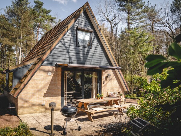 Una pequeña cabaña de madera con forma triangular, fachada de ladrillo, una terraza de madera y una parrilla.