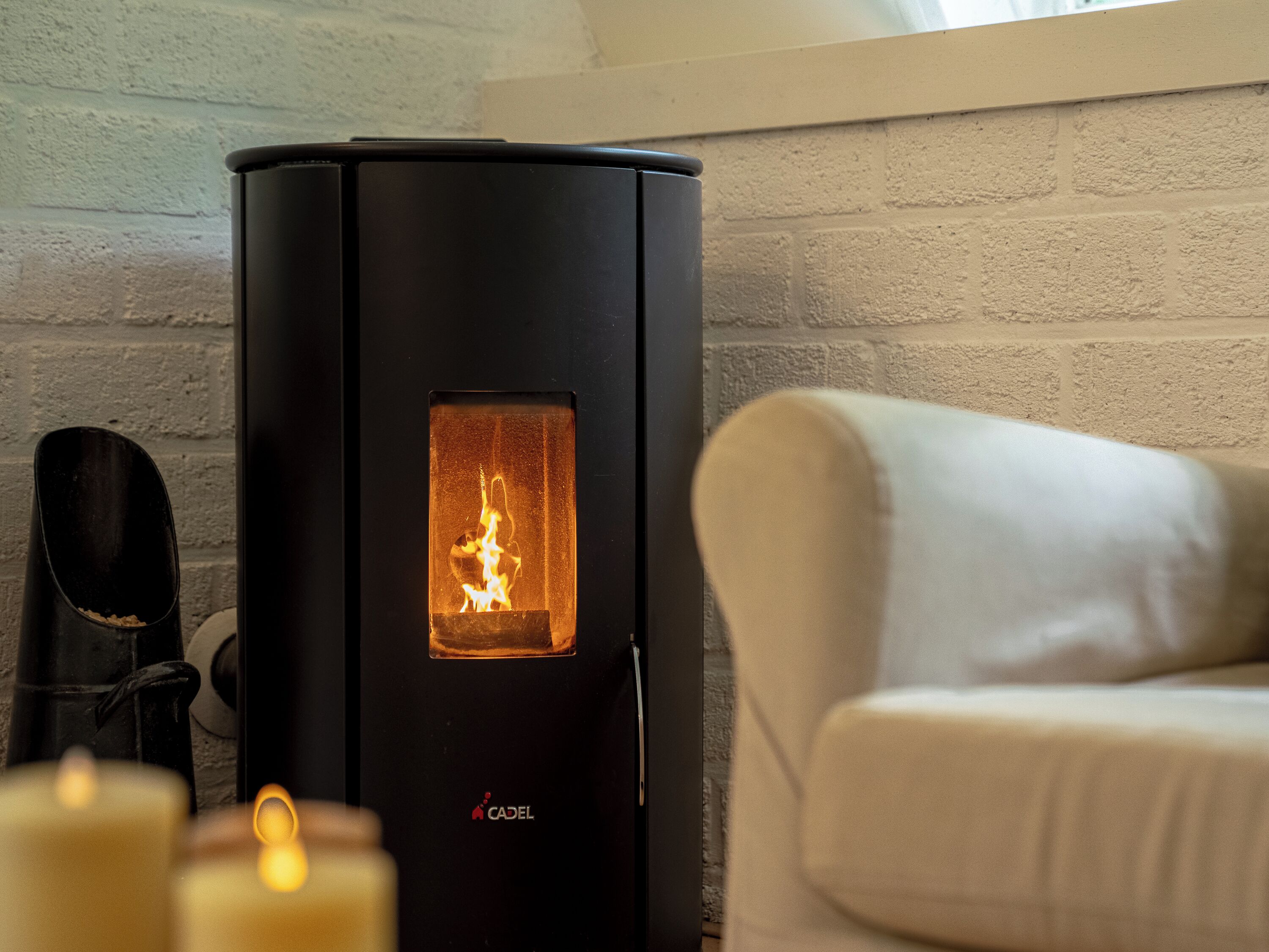 Un poêle à bois noir avec une flamme visible, un porte-chaussures noir et un fauteuil beige dans une pièce dont un mur est en briques blanches.