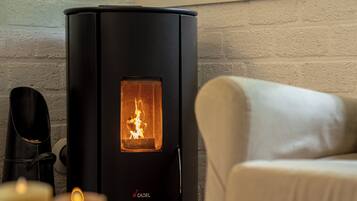 Un poêle à bois noir avec une flamme visible, un porte-chaussures noir et un fauteuil beige dans une pièce dont un mur est en briques blanches.