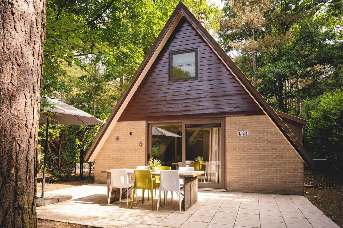 A small, triangular wooden cabin with a patio area, outdoor seating, and a door leading to a covered entrance.
