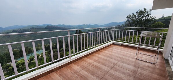 Un balcón con piso de madera y barandilla blanca, con vista a un paisaje verde con colinas y árboles.