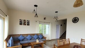 A spacious living room with a wooden sofa, a coffee table, and a dining table with chairs. There are hanging lights and a clock on the wall.