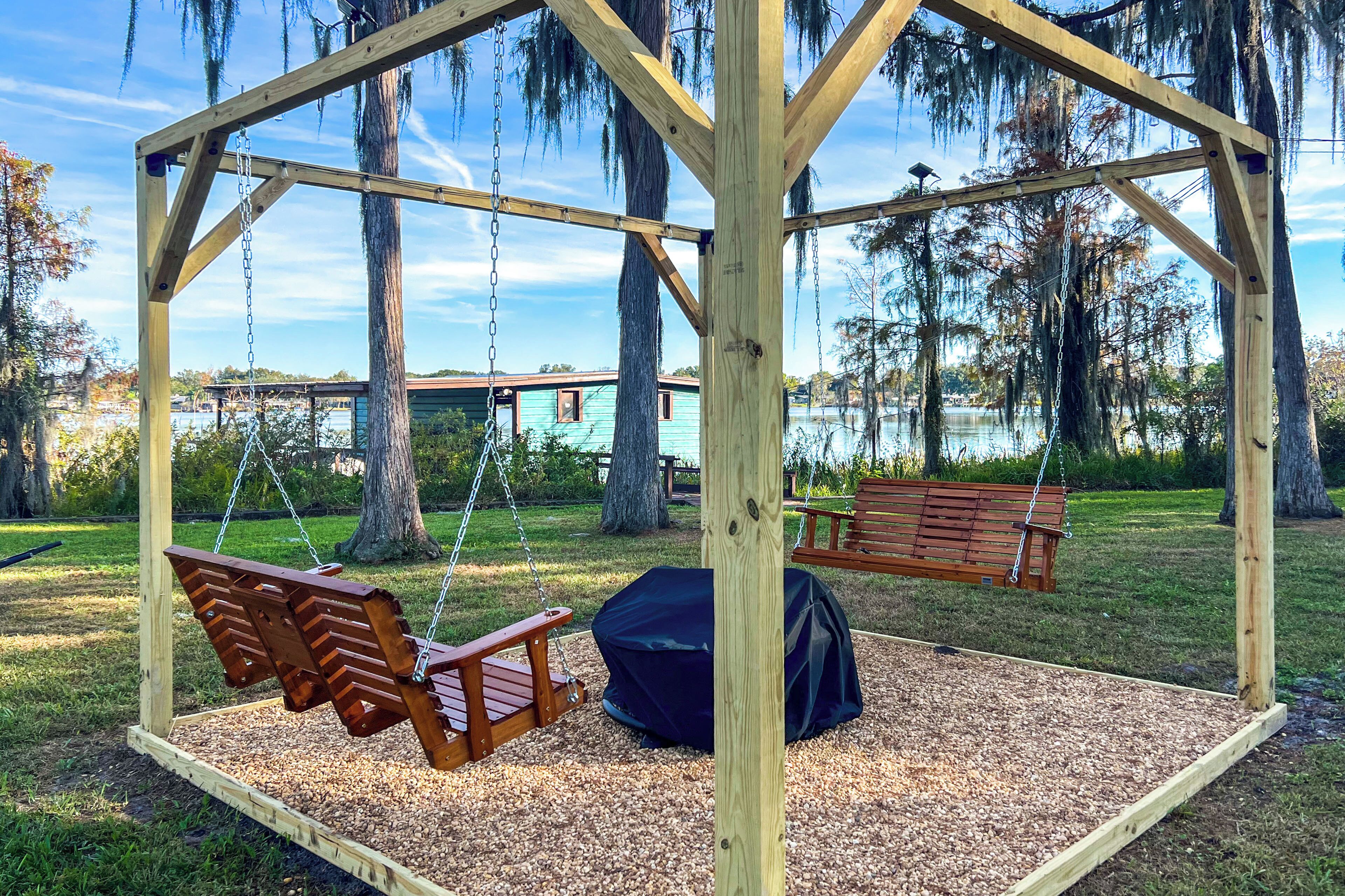 A wooden swing set with benches and a covered seating area, surrounded by trees and a body of water in the background.