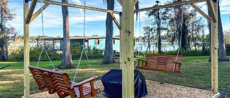 A wooden swing set with benches and a covered seating area, surrounded by trees and a body of water in the background.
