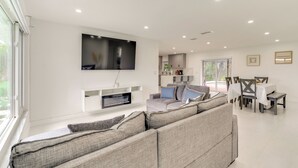 A modern living room with a large flat-screen TV mounted on the wall, a grey sectional sofa, and a dining area with a table and chairs. - Swim, Golf & Explore: Pet-friendly Cape Coral Home (Cape Coral)