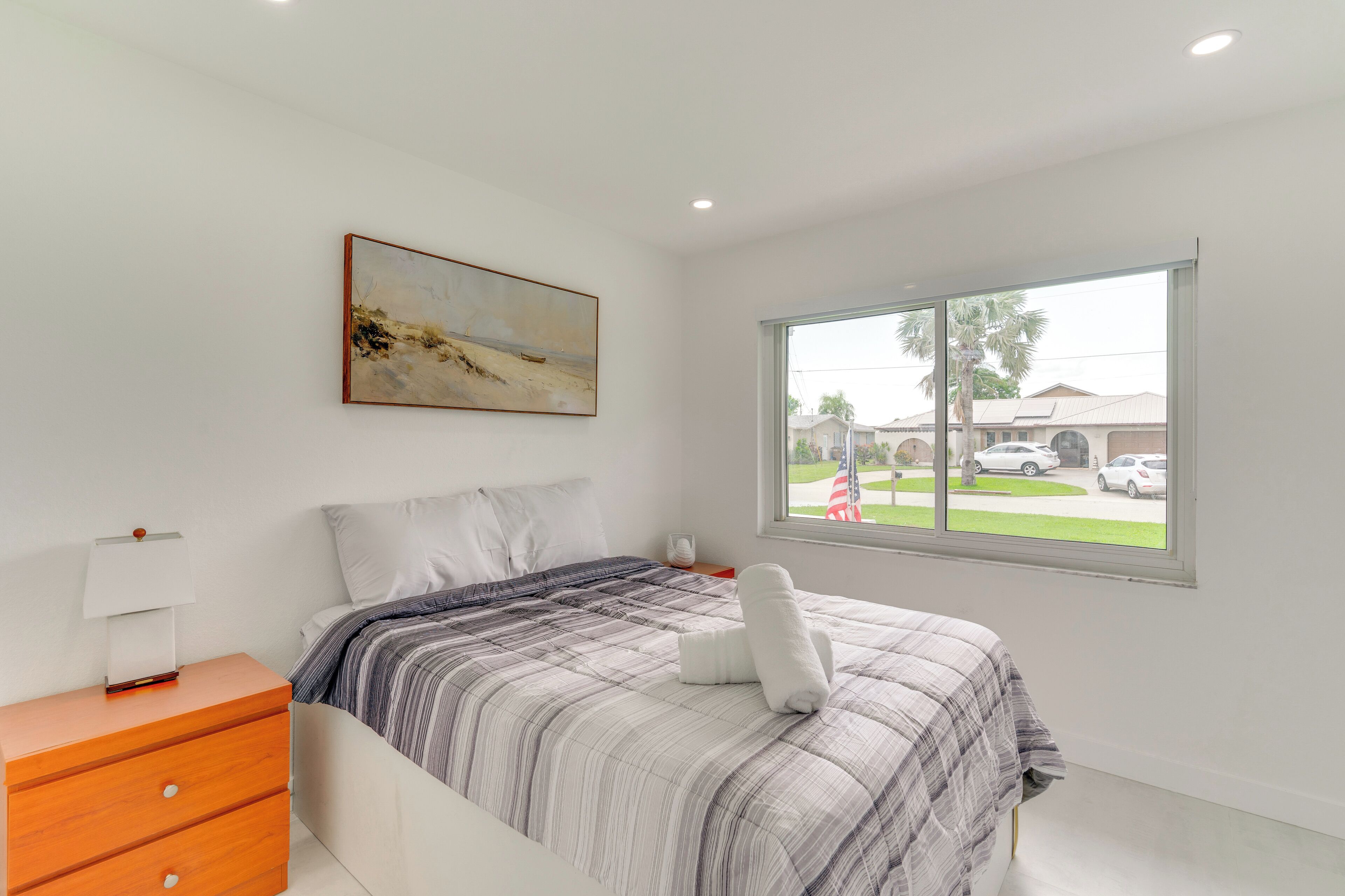 Un dormitorio con cama, mesita de noche, un cuadro y ventana con vistas al exterior.