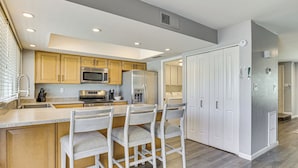 A modern kitchen with wooden cabinets, stainless steel appliances, and a bar area with white chairs.