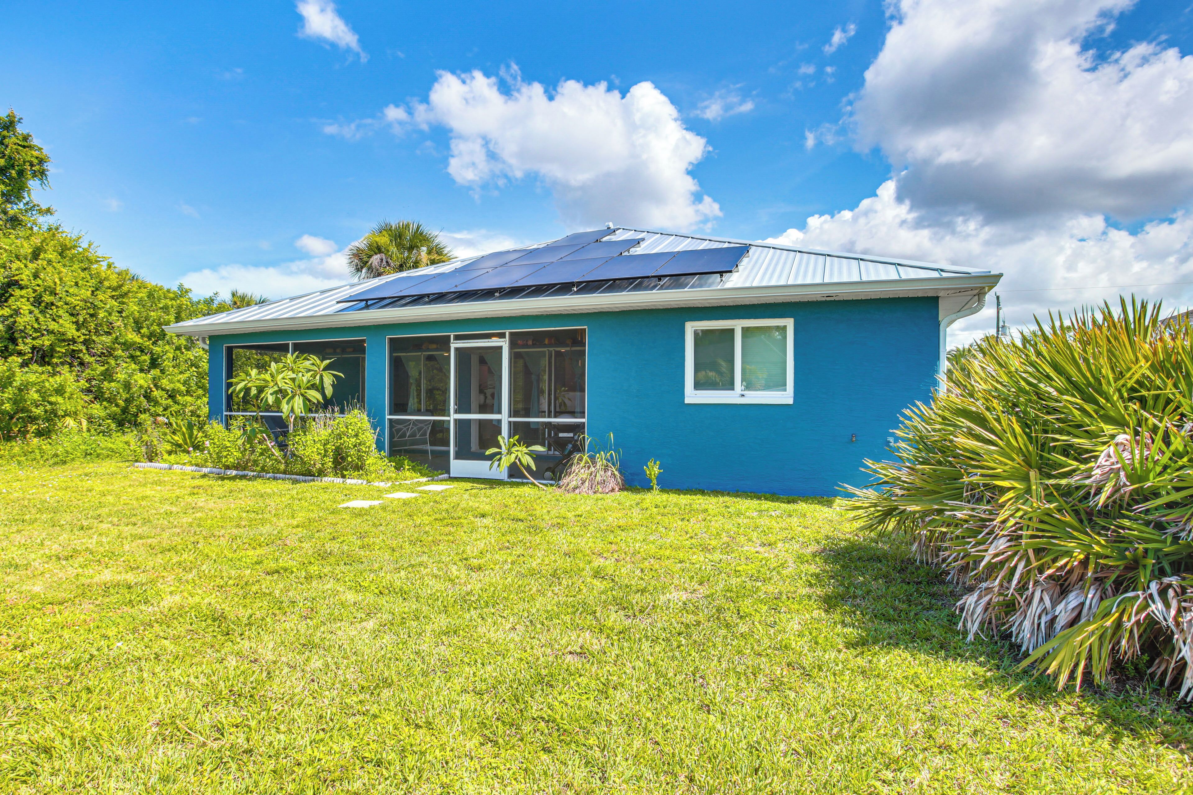 Una casa de un solo piso con fachada azul, techo con paneles solares y un patio cubierto de césped y plantas tropicales.
