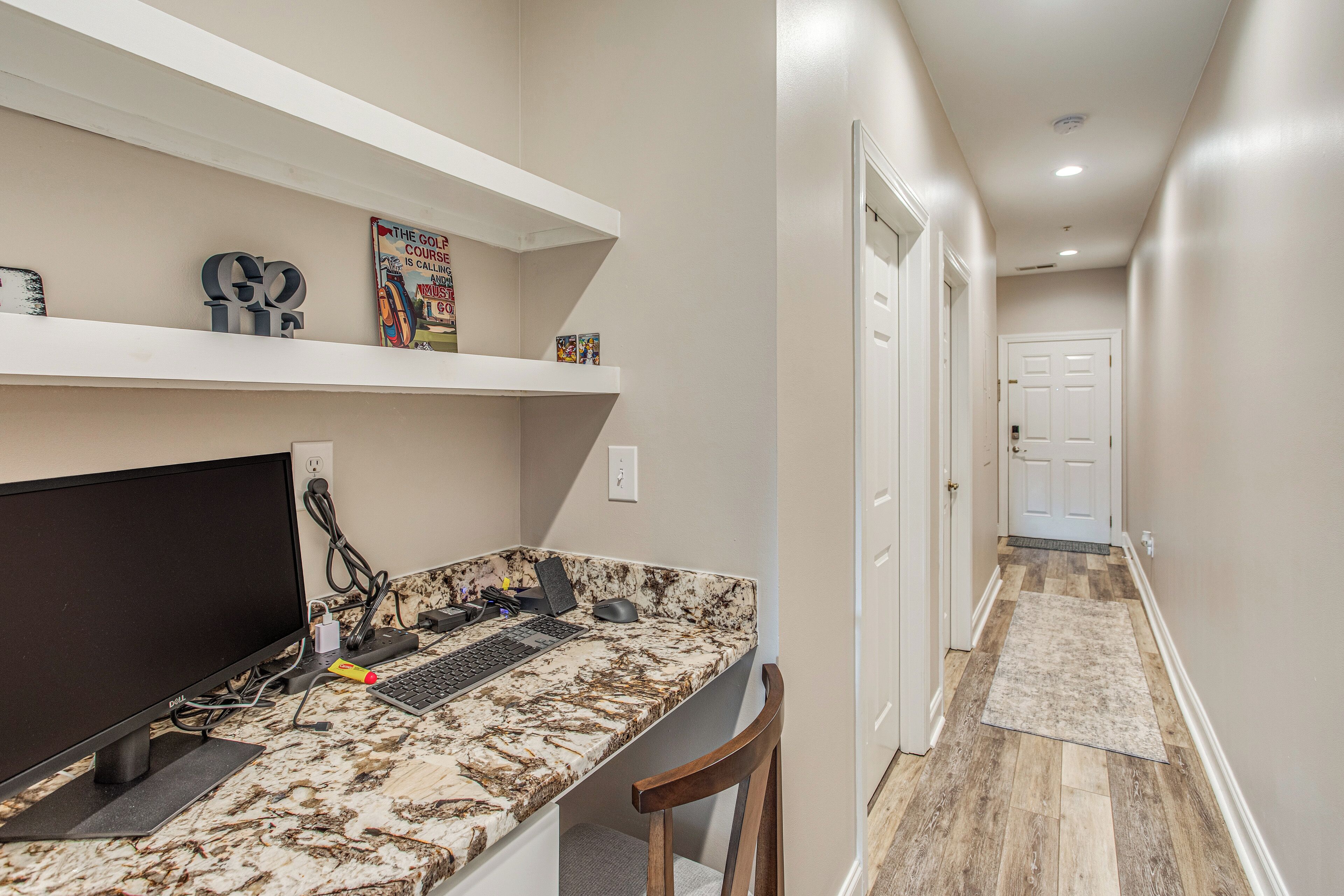 A home office with a desktop computer, shelves with decorative items, and a hallway leading to a door.