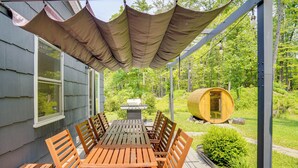 Een overdekt terras met een houten tafel en stoelen, een barbecue en een houten vat.