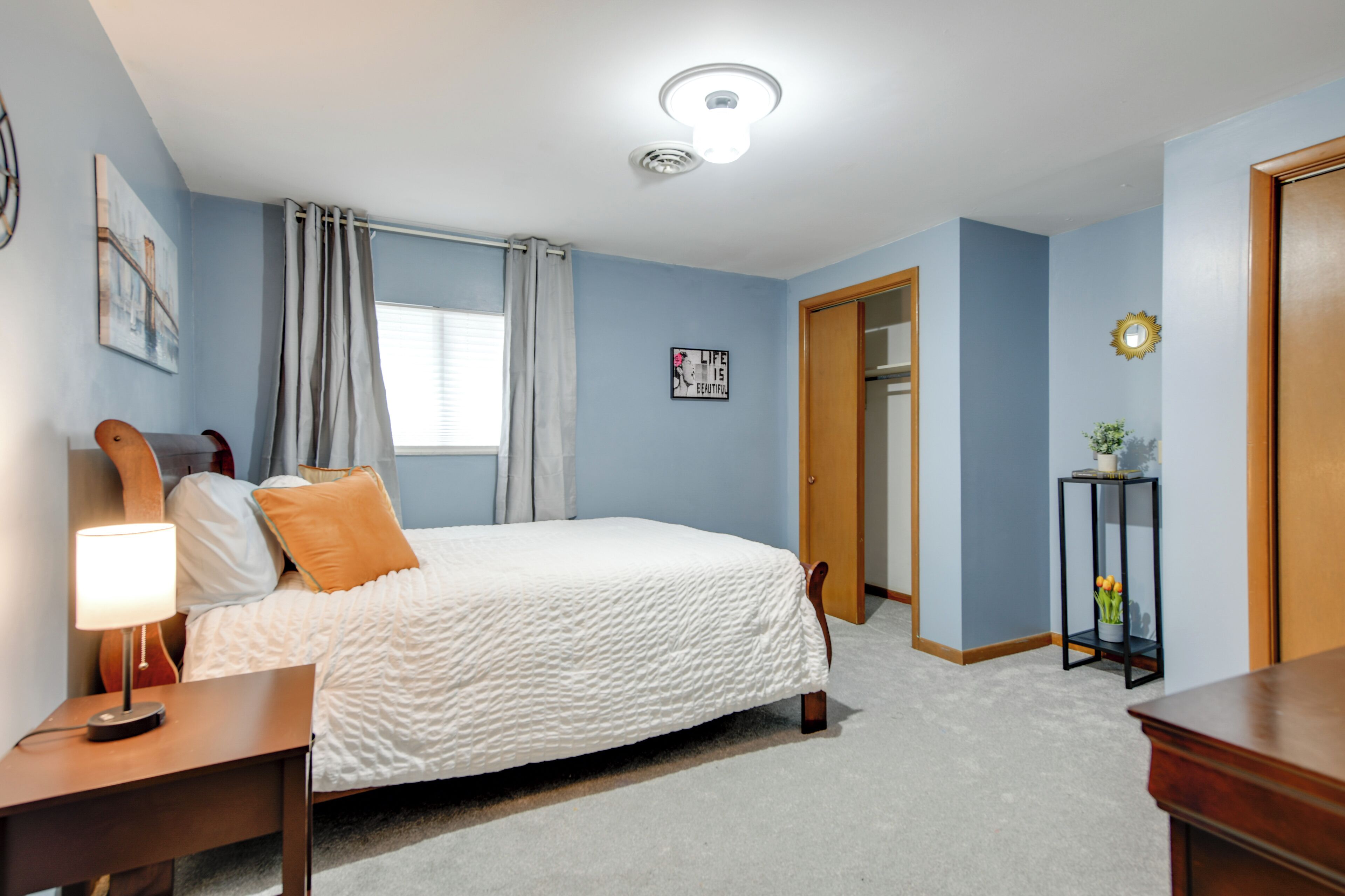Quarto com cama, mesa de cabeceira, luminária, janela com cortinas, porta e relógio na parede.