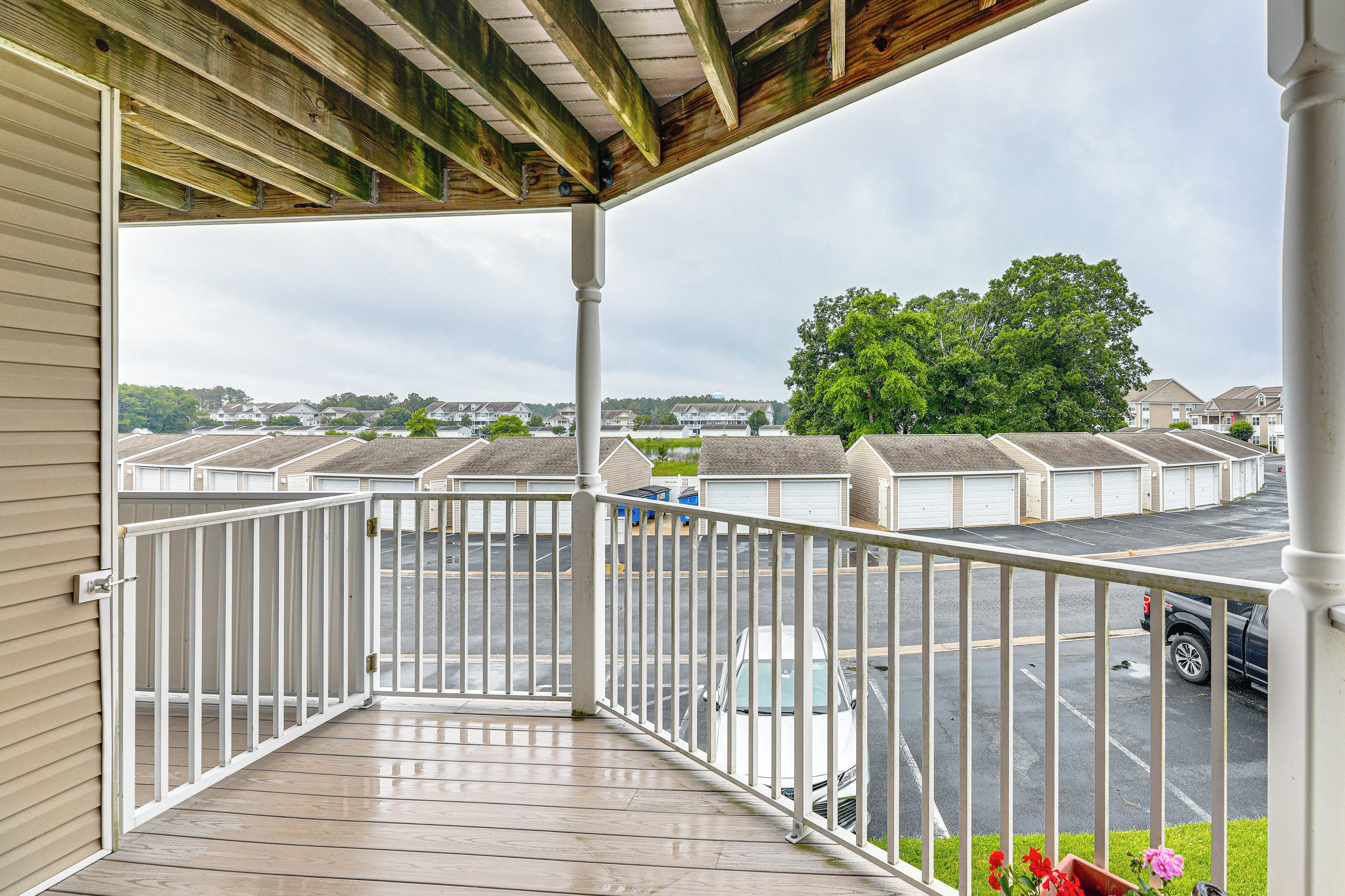 A covered outdoor area with a wooden deck, white railing, and a view of a parking lot with several cars and a row of houses in the background.