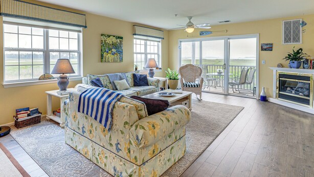 A living room with a floral sofa, a dining area with a table and chairs, and a view through a glass door to the outdoors. - Swim, Fish & Explore: Marsh-front Gem in Delaware! (Ocean View)