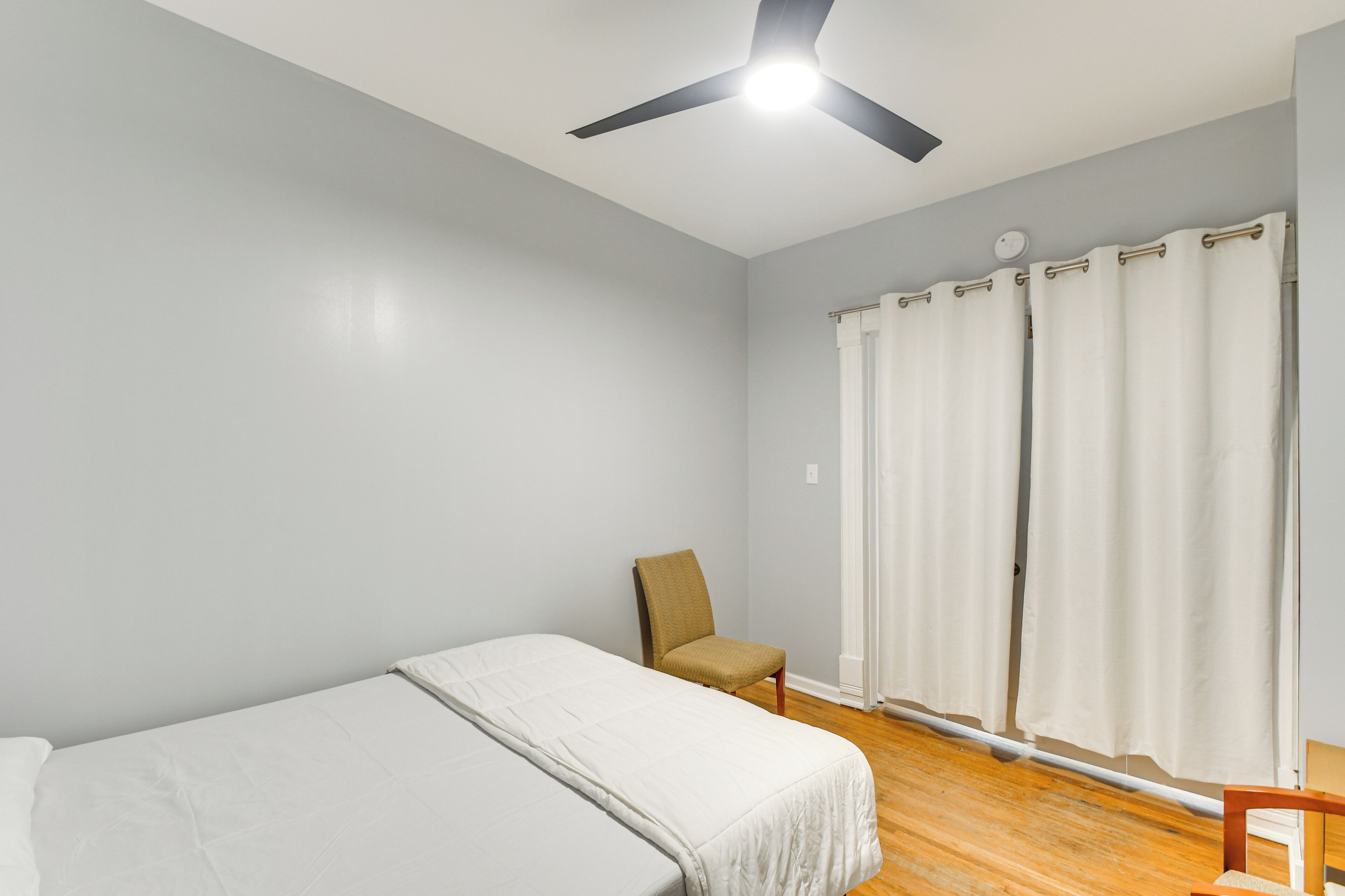 Quarto com cama, poltrona, ventilador de teto e janela com cortinas.