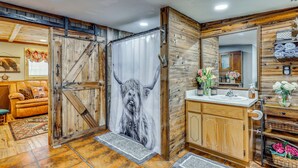 A bathroom with a wooden wall, a shower curtain with a bull's head design, a sink with a mirror, and a cabinet with toiletries. - Quiet Eagar Retreat Near Lakes, Hiking & Hunting! (Eagar)